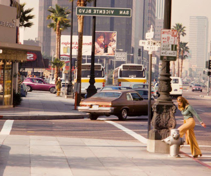 Image of Beverly Hills in the 1970s with period cars turning a busy street and a young woman crossing the street.