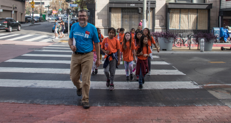 学生s in orange shirts cross the street with an adult tour guide.